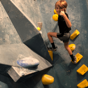 A boy on an obstacle at a rock climbing gym gives a thumbs up.