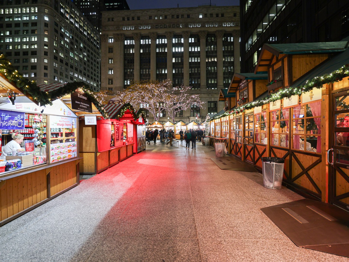 A wide, uncrowded aisle between rows of lit wooden holiday huts trimmed with garlands, with a few visitors near trees wrapped in white lights
