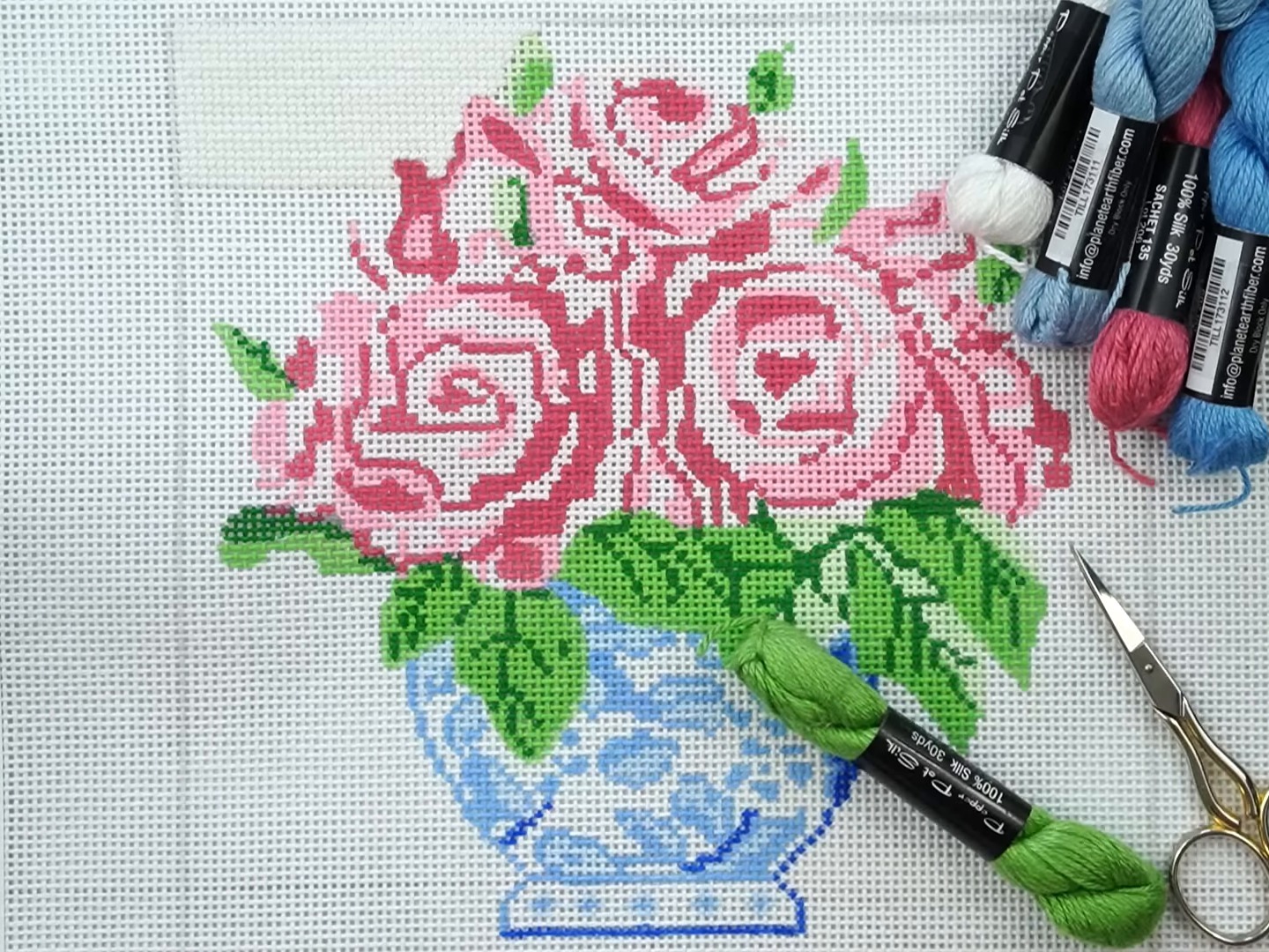 A painted needlepoint canvas of pink roses in a blue and white vase lies on a table beside skeins of pink, blue, white and green thread and small gold embroidery scissors