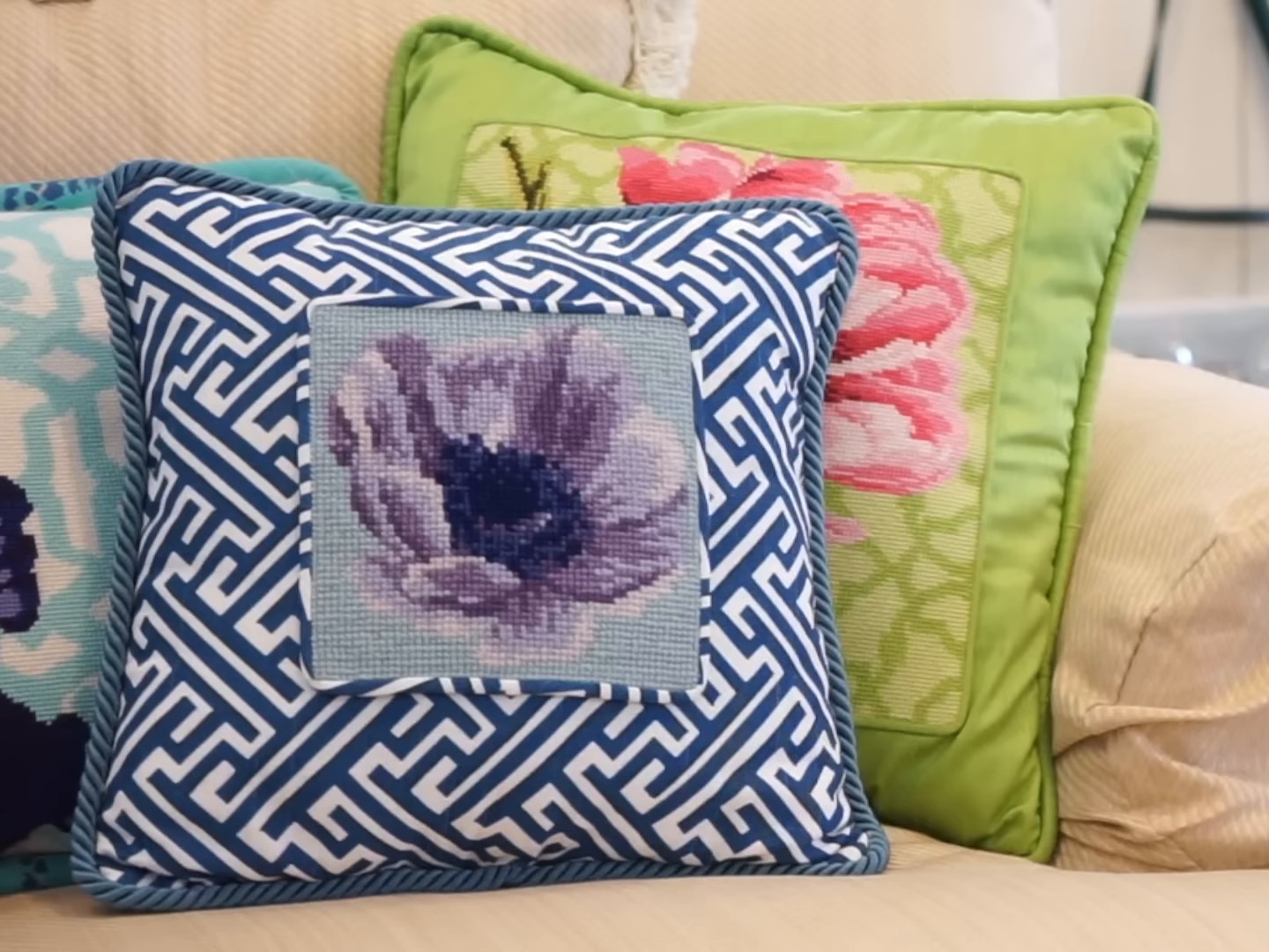 Finished needlepoint pillows on a cream sofa, one with a purple flower framed by a blue and white geometric border and one with pink tulips on green