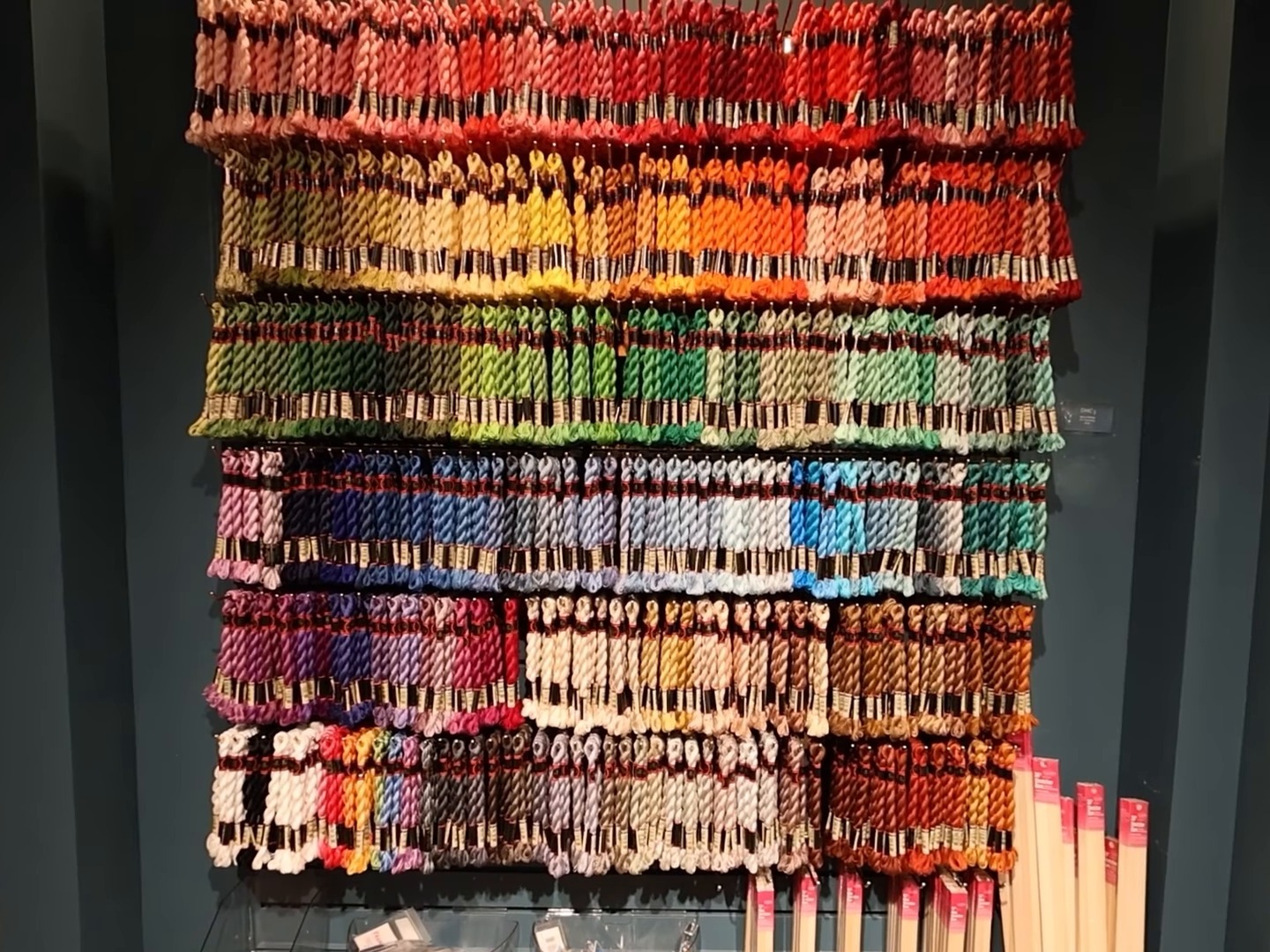 A dark teal shop wall hung with rows of needlepoint thread arranged like a rainbow, from reds and oranges through greens, blues and purples