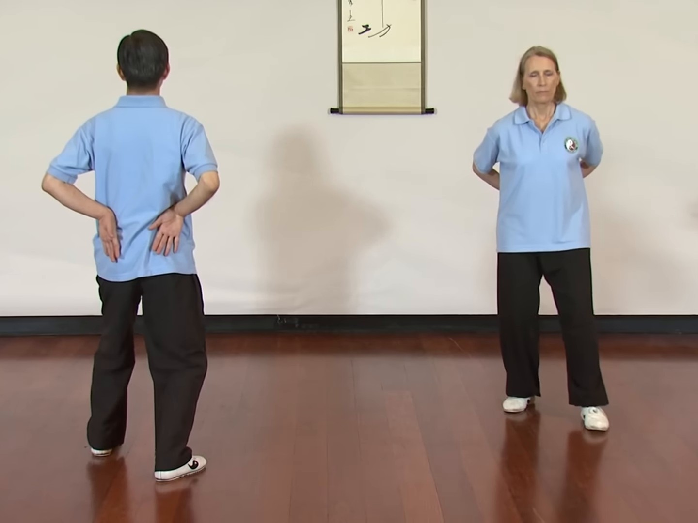 Dr Paul Lam, seen from behind, and a fellow teacher facing the camera stand on a wooden studio floor with the backs of their wrists resting on their lower backs