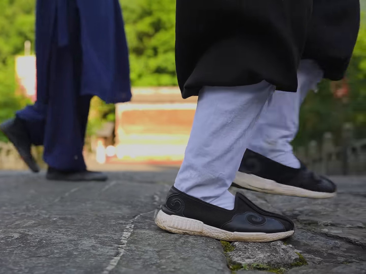 Close view of two walkers' feet in black cloth shoes on old stone paving, one foot planted flat while the back heel lifts