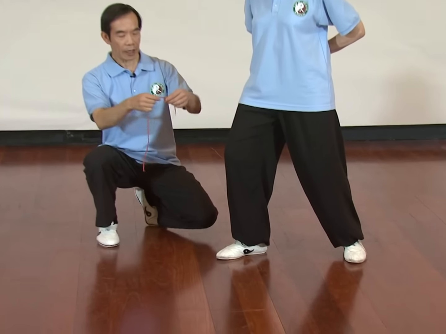 Dr Paul Lam crouches beside a teacher standing in a shallow stance on a wooden floor and holds up a thin red string to show the line from knee to toe