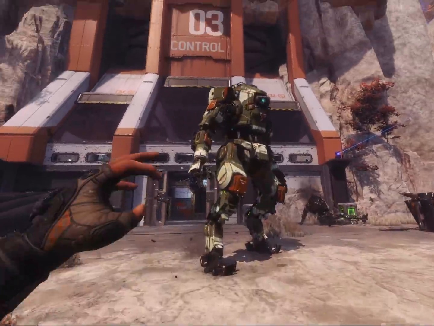 First person view of a gloved hand reaching toward a giant green and white Titan robot walking out of a hangar marked 03 Control