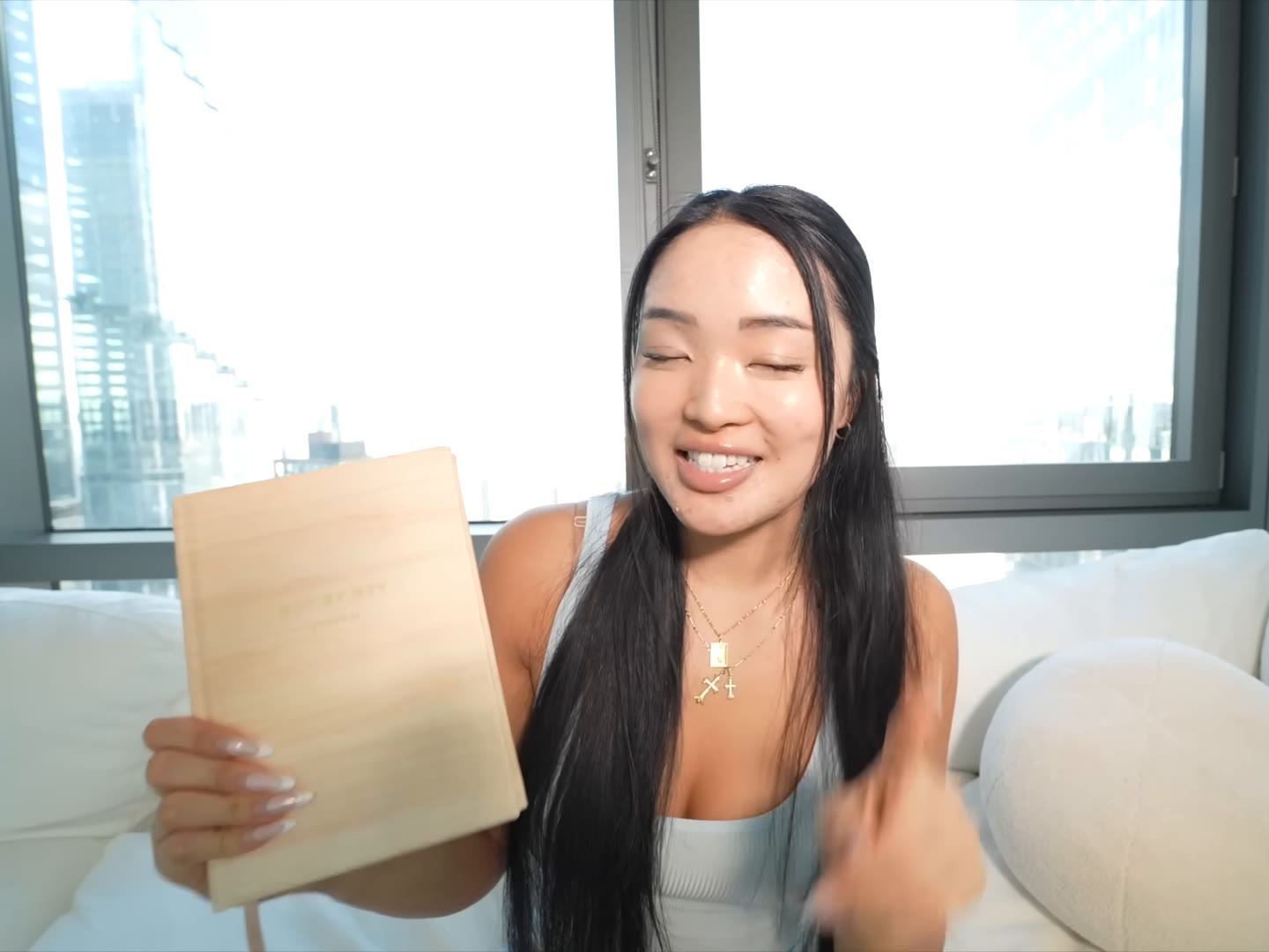 Smiling woman on a white sofa holding up a tan notebook in front of a city window