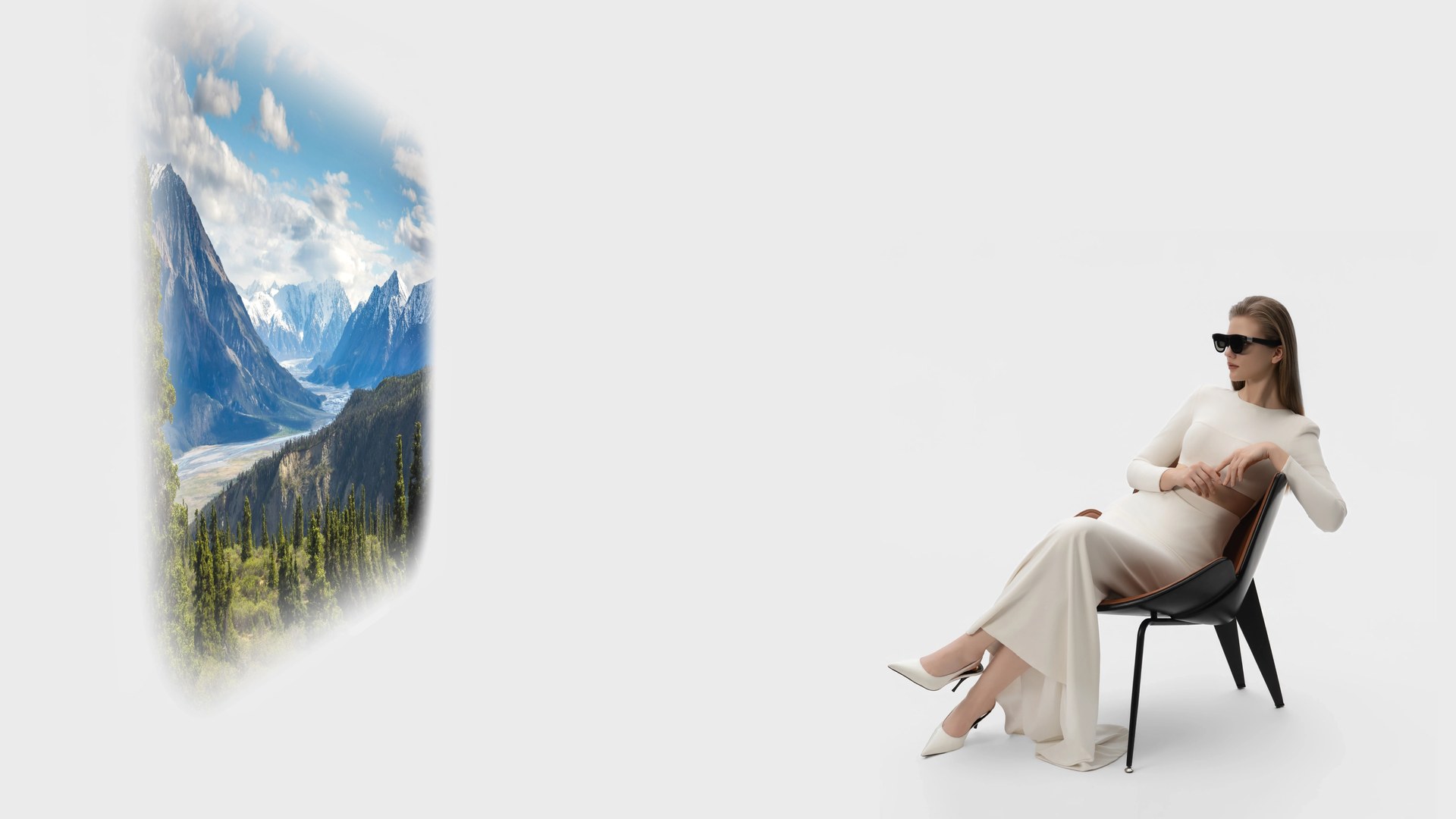 A woman in a long cream dress relaxes in a lounge chair wearing XREAL AURA glasses while a large virtual window shows snowy mountains and a pine forest