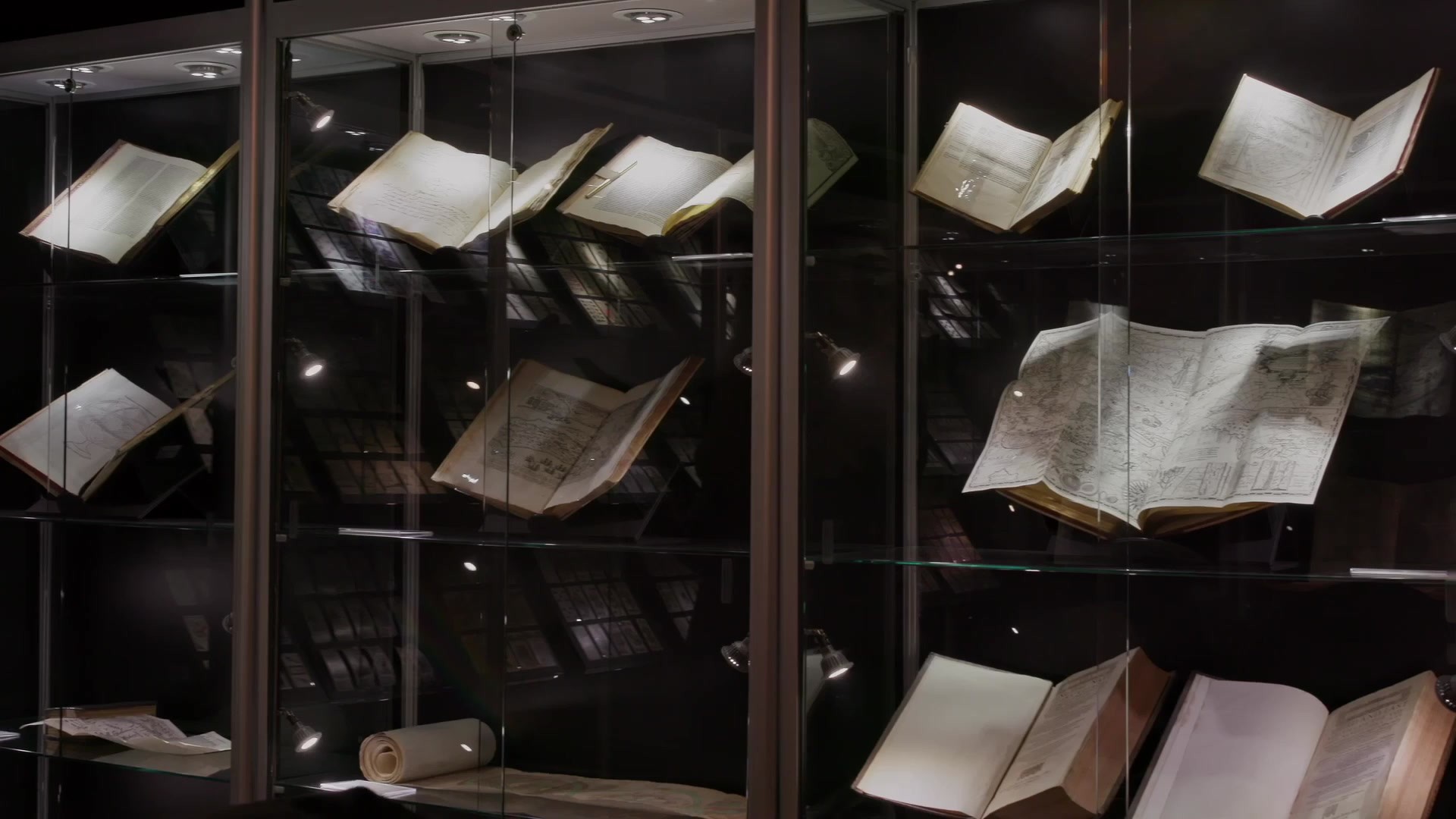Open antique books and a scroll rest on glass shelves inside softly lit display cases in a dark room