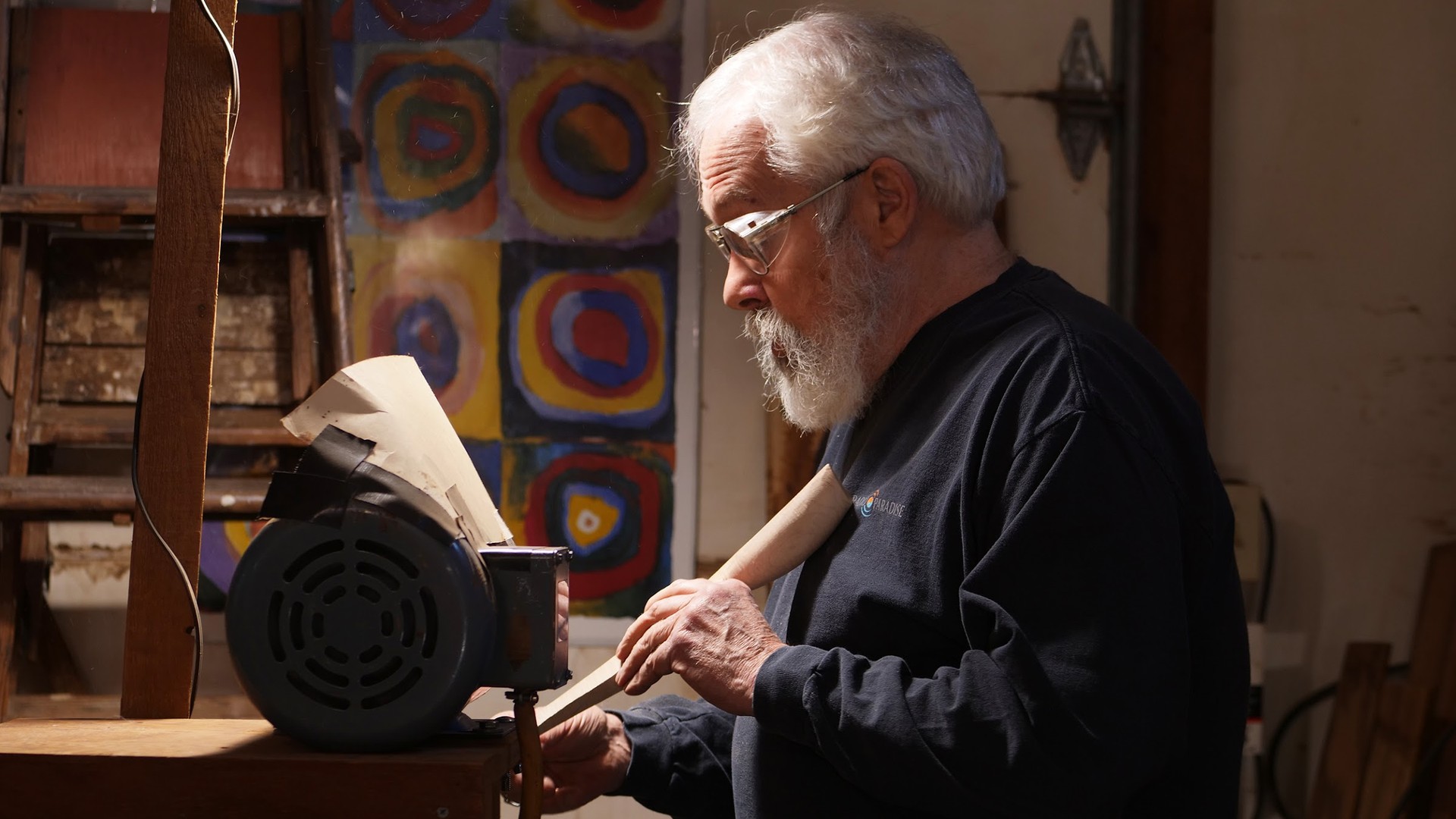 A white bearded wood artist in glasses and a dark sweatshirt shapes a wooden spoon handle at a sanding machine in his workshop