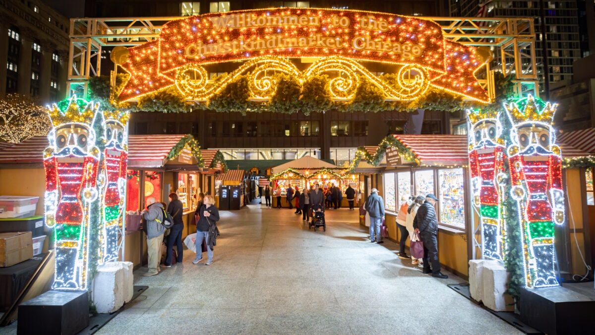 A glowing arch of lights reading Willkommen to the Christkindlmarket Chicago, flanked by lit nutcracker figures, opens onto an aisle of wooden holiday stalls with shoppers