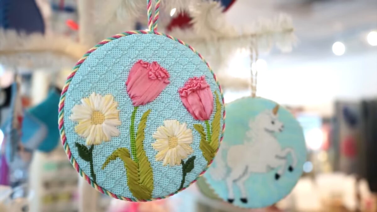A round needlepoint ornament stitched with pink tulips and white daisies on a pale blue background hangs in a softly blurred craft shop