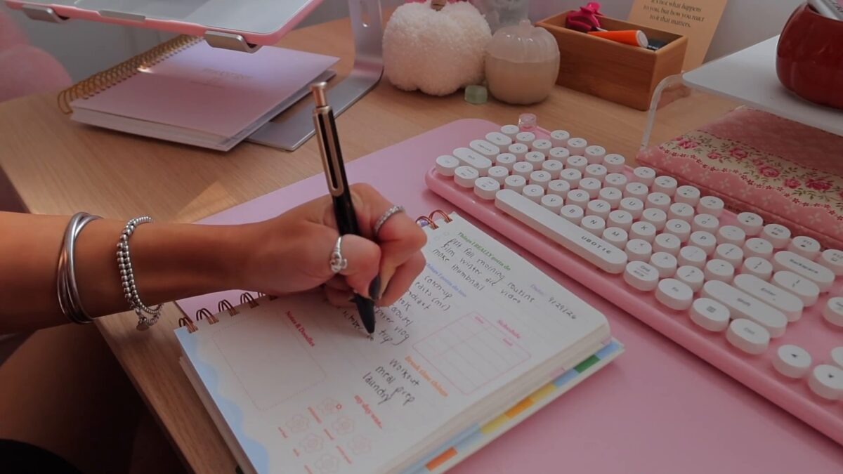 Hand writing a winter arc to-do list in a spiral planner beside a pink keyboard and white pumpkins