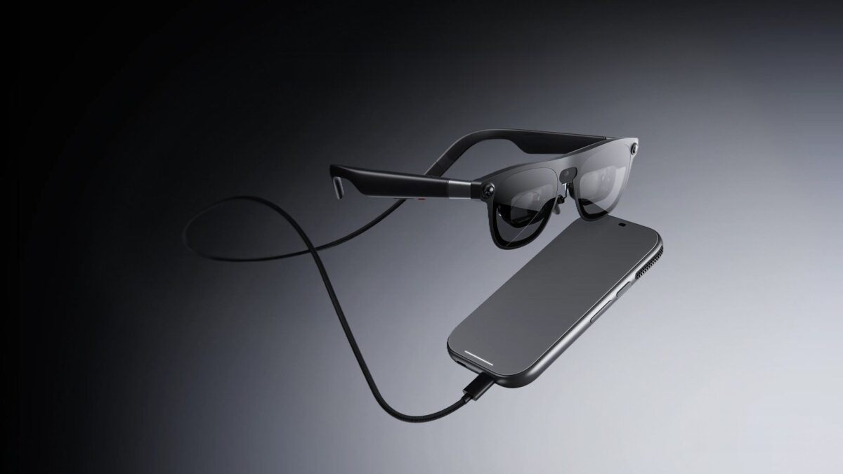 XREAL AURA glasses in black, connected by a cable to a slim black compute puck, on a soft gray gradient background