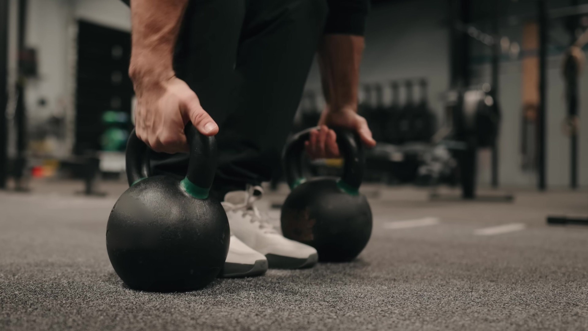 A close view of an athlete gripping two black kettlebells on a dark gym floor before a farmers carry