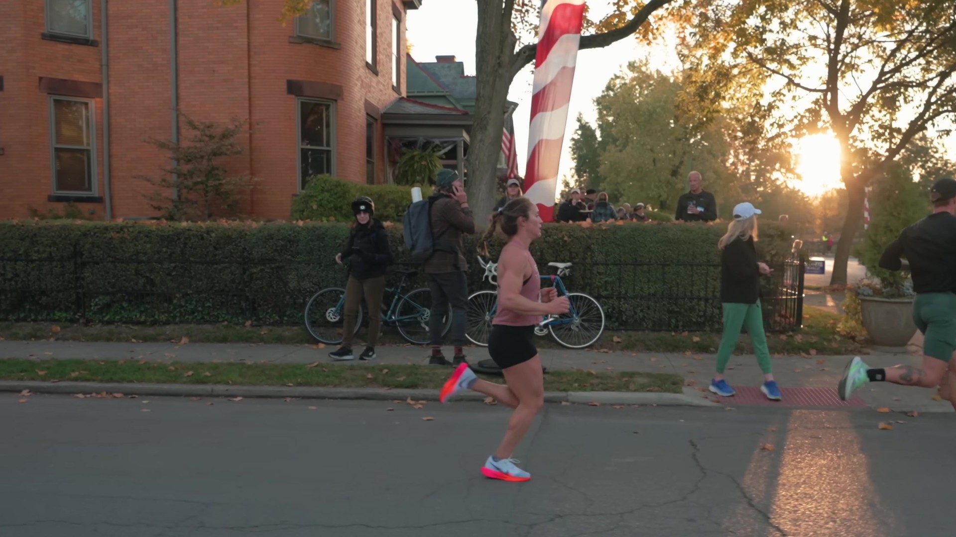 Runners pass brick houses and autumn trees on a city street as the low sun glows through the branches