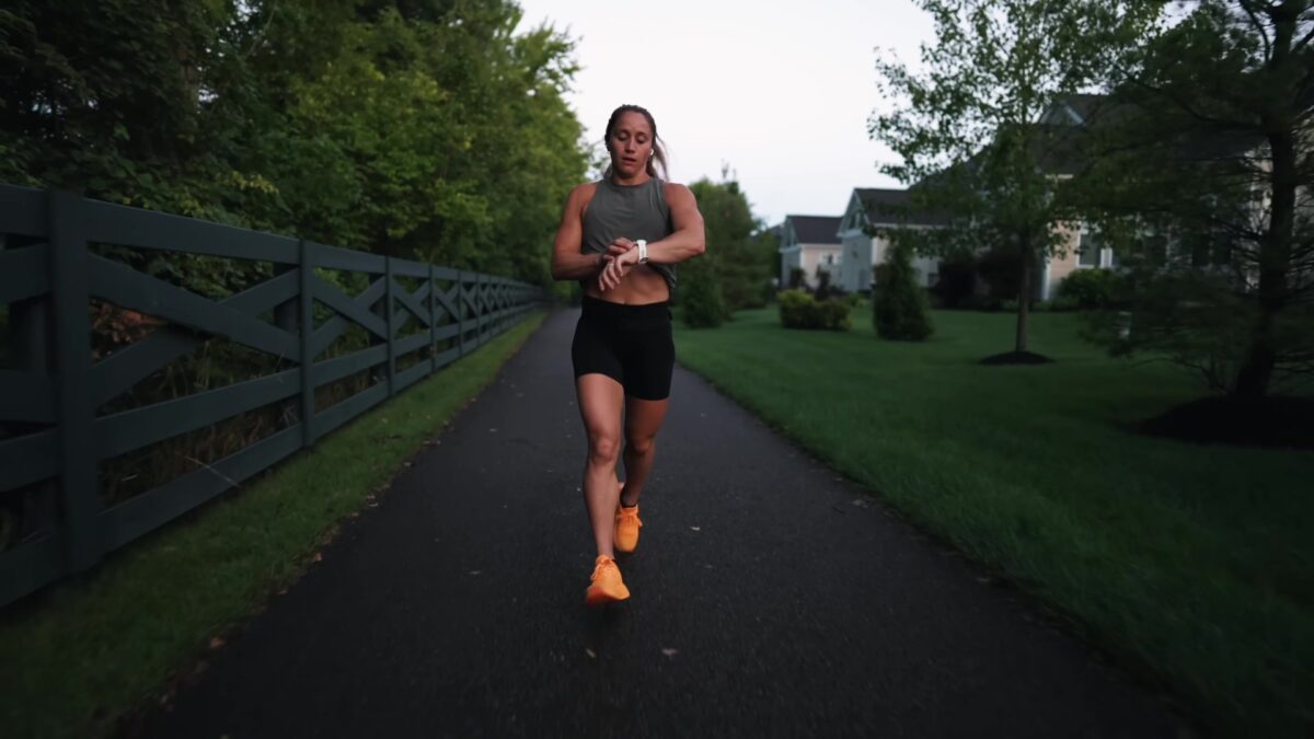 A woman in a gray tank top and orange running shoes runs along a quiet tree lined path beside a dark wooden fence at dusk
