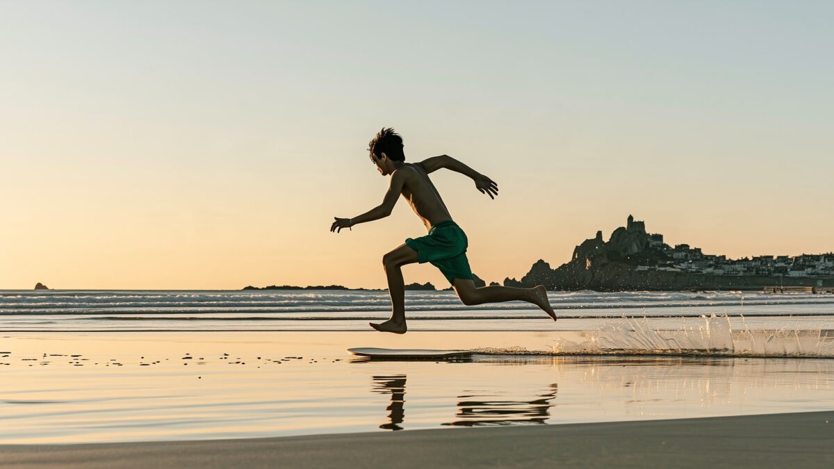 A skimboarder running and jumping onto his board on the wet sand at sunset