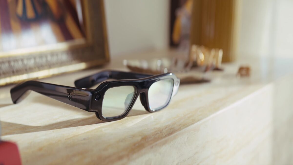 A pair of chunky black Snap Specs augmented reality glasses resting on a cream marble mantel in warm afternoon light, beside a gilded picture frame