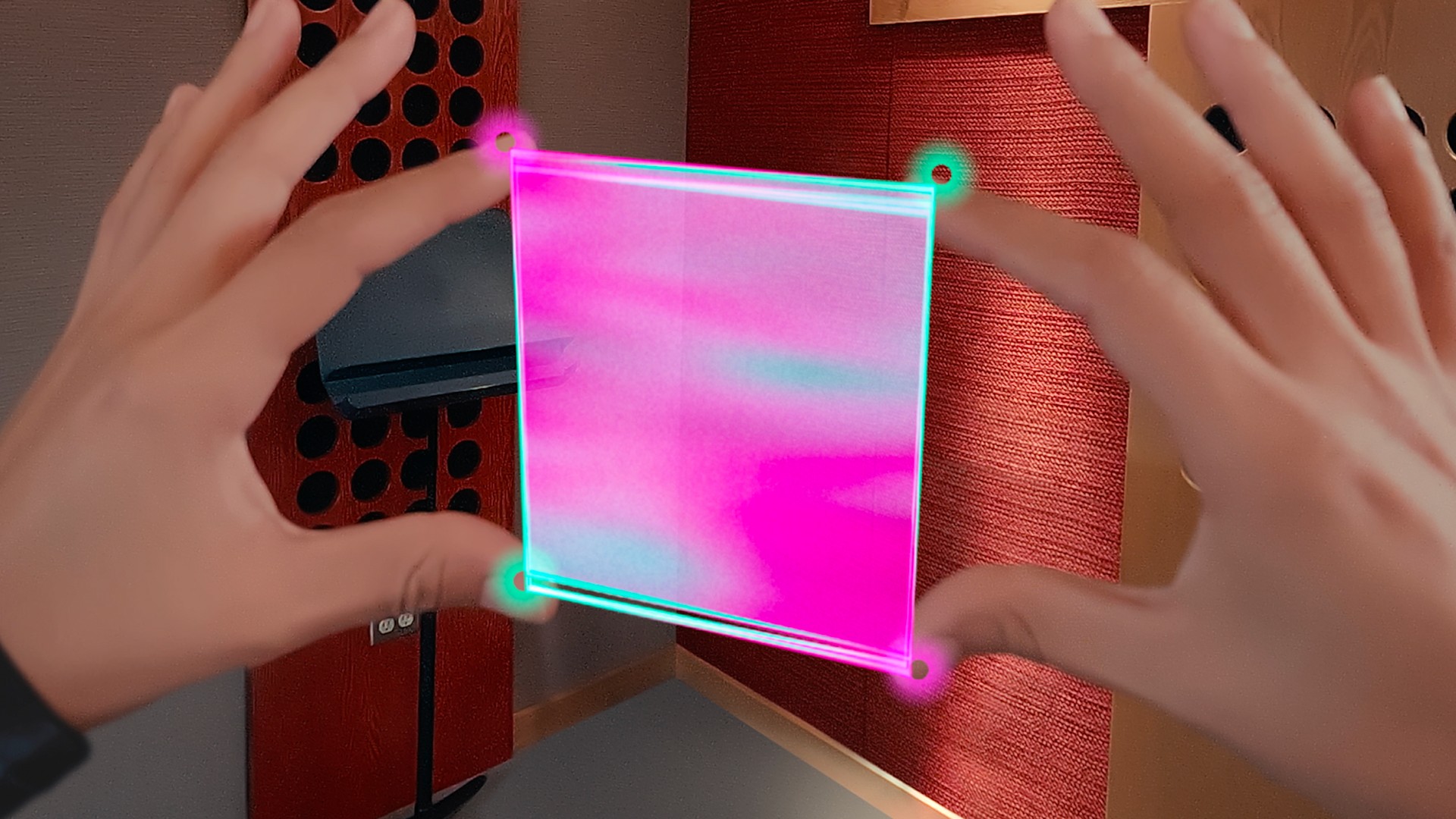 Two hands frame a glowing pink and turquoise virtual panel in the air inside a small music studio with red wood acoustic panels and a music stand