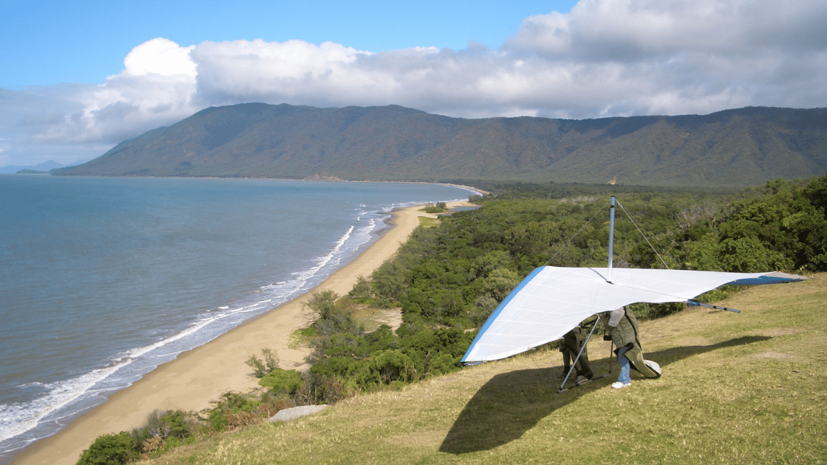 A man and his hang glider sit on a hill overlooking the ocean.